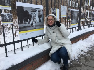 kobieta w średnim wieku, w zimowym stroju, kuca przy ogrodzeniu, na którym wiszą plansze ze zdjęciami. Wkoło leży śnieg. 