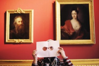 Kobieta spoglądająca przez wycięte otwory w kartce papieru. Kobieta stoi na tle portretów zawieszonych na ścianie w Pałacu na Wyspie.