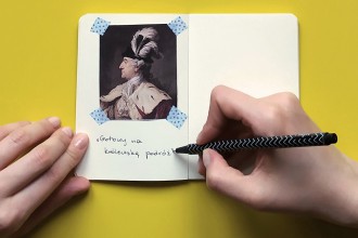 Na żółtym stole leży notatnik z wklejoną miniaturą portretu króla Stanisława Augusta. Lewa dłoń podtrzymuje brzeg kartki, prawa trzyma długopis.