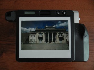 Na drewnianym blacie leży aparat do wykonywania natychmiastowych odbitek. Na nim położone zdjęcie Pałacu na Wyspie.