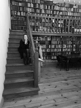Czarno-białe zdjęcie. Dr Akvilė Naudžiūnienė siedzi na drewnianych schodach, w tle półki z książkami.