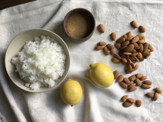 Składniki potrzebne do przygotowania zupy migdałowej ułożone na kremowej serwecie: (od lewej) miska z ugotowanym ryżem, dwie cytryny, miseczka z cukrem brązowym, migdały.