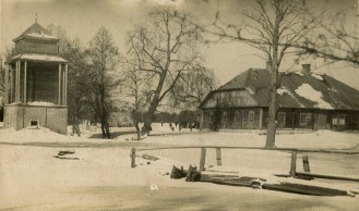 wiejski krajobraz, z lewej drewniana dzwonnica, z prawej duży drewniany dom, pomiędzy bezlistne drzewa. Wszystko zaśnieżone