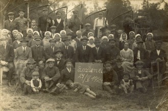 Fotografia w sepii. Grupa kilkudziesięciu mężczyzn, kobiet i dzieci pozuje na tle krajobrazu. Osoby w pierwszym rzędzie trzymają tabliczkę z napisem: "Łazienki 27/V. 1924"