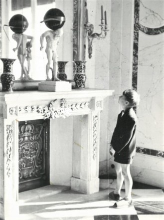 Czarno-biała fotografia. Wnętrze Jadalni w Pałacu na Wyspie, chłopiec spogląda na stojący przed nim, na kominku zegar w formie rzeźby mężczyzny dźwigającego kulę.
