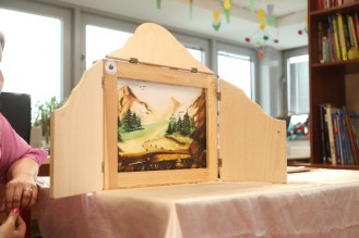 na zdjęciu widać wnętrze pokoju w którym na pokrytym bladoróżowym obrusem stole postawiono rozkładaną drewnianą ramę w której widać obraz przedstawiający  las