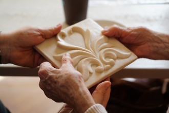 Na zdjęciu widać dłonie należące do starszej osoby, trzymające oraz poznające poprzez dotyk kwadratową płytkę z jasnego kamienia z płaskorzeźbionym ornamentem.